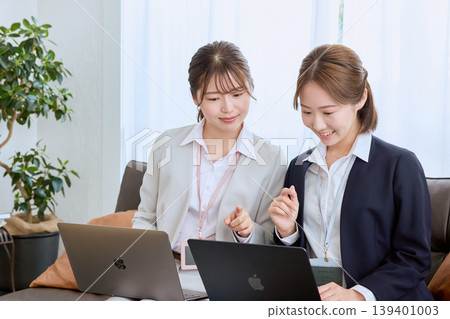 Two young female business professionals are having a meeting while looking at their laptop screens – a scene of a business negotiation in an office. Two young female business professionals are having a meeting while looking at their laptop screens – a scene of a business negotiation in an office. 139401003