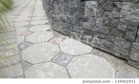 High angle view shows curved dark stone wall beside hexagonal pavement tiles. Grey masonry, red accents create geometric patterns. This durable surface suits architecture, landscaping, urban design. High angle view shows curved dark stone wall beside hexagonal pavement tiles. Grey masonry, red accents create geometric patterns. This durable surface suits architecture, landscaping, urban design. 139402051