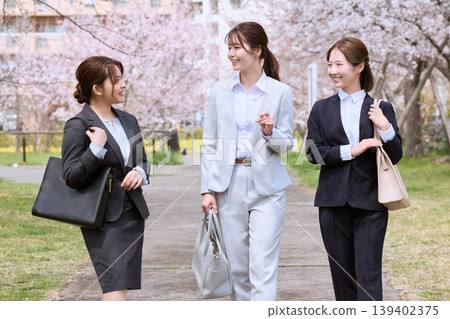 Three young female new employees walking under a row of cherry trees; job seekers in suits with smiles; an image of a spring induction ceremony. 139402375