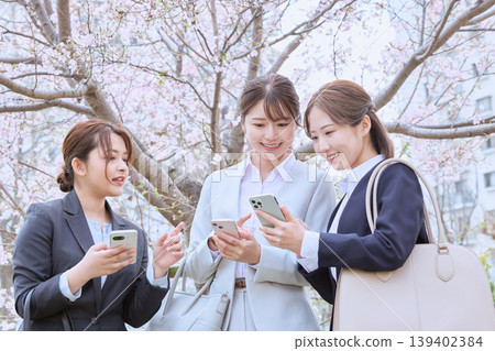 A spring scene: three young female business professionals/new employees chatting while looking at their smartphones under the cherry blossoms. A spring scene: three young female business professionals/new employees chatting while looking at their smartphones under the cherry blossoms. 139402384