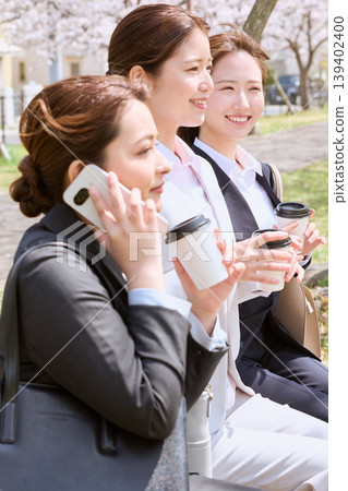 三位年輕的職業女性在一排櫻桃樹下一邊聊天一邊看著智慧型手機;享受著春天戶外的咖啡時光。 三位年輕的職業女性在一排櫻桃樹下一邊聊天一邊看著智慧型手機;享受著春天戶外的咖啡時光。 139402400