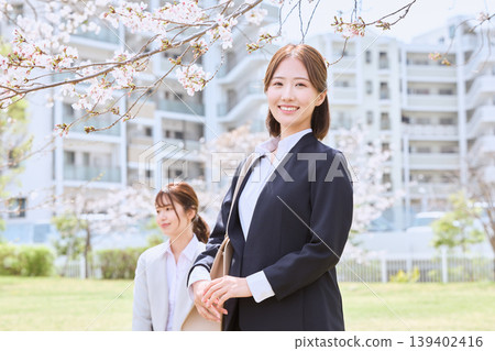 An image of a spring company induction ceremony, with young women in suits and female university students smiling as they look for jobs on a campus where cherry blossoms are in bloom. 139402416