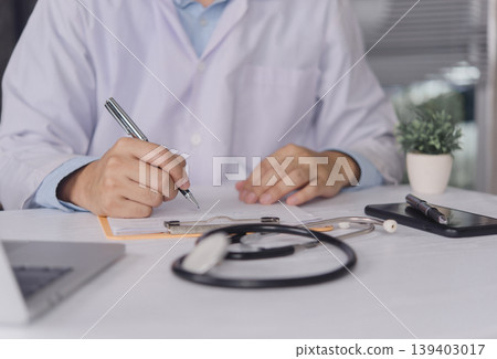 Close up doctor hand writing prescription on clipboard with stethoscope on desk. Professional physician filling medical report and insurance form in hospital clinic. Healthcare administrative concept. Close up doctor hand writing prescription on clipboard with stethoscope on desk. Professional physician filling medical report and insurance form in hospital clinic. Healthcare administrative concept. 139403017