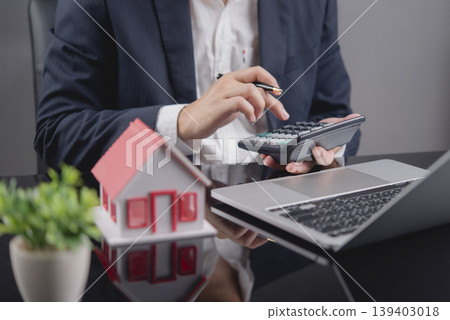Businessman using calculator for calculating finance on table with house model. Real estate, tax, and property investment concept. Businessman using calculator for calculating finance on table with house model. Real estate, tax, and property investment concept. 139403018