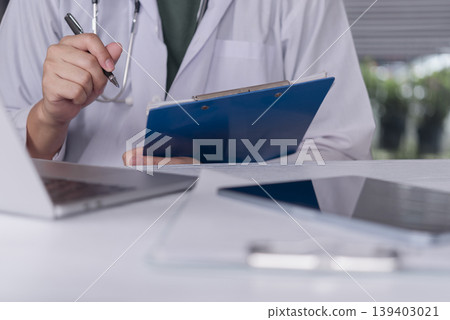 Close up doctor hand writing medical report on clipboard with digital tablet on desk. Professional physician working with paperwork and electronic health record technology in hospital office. Close up doctor hand writing medical report on clipboard with digital tablet on desk. Professional physician working with paperwork and electronic health record technology in hospital office. 139403021