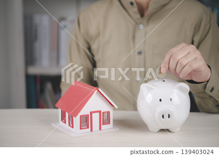 Man putting coin into white piggy bank near house model. Concept of saving money for buying new home, real estate investment, and mortgage financial planning. Man putting coin into white piggy bank near house model. Concept of saving money for buying new home, real estate investment, and mortgage financial planning. 139403024