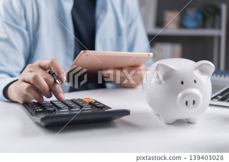 Man using calculator and holding digital tablet for financial planning. Budgeting, tax return calculation and savings concept with white piggy bank on desk. Man using calculator and holding digital tablet for financial planning. Budgeting, tax return calculation and savings concept with white piggy bank on desk. 139403028