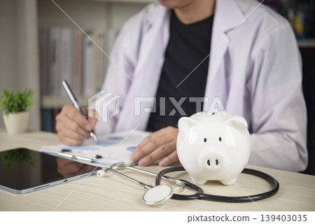 Doctor with stethoscope and white piggy bank on desk, concepts of healthcare costs, medical insurance planning, and saving money for health treatment expenses. Doctor with stethoscope and white piggy bank on desk, concepts of healthcare costs, medical insurance planning, and saving money for health treatment expenses. 139403035