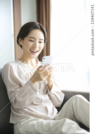 一位女士看著自己的智慧型手機,臉上露出笑容,享受著在家中的輕鬆時光。 一位女士看著自己的智慧型手機,臉上露出笑容,享受著在家中的輕鬆時光。 139407141
