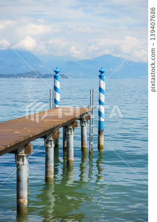 Wooden pier extending into Lake Garda with decorative blue striped poles, calm water and mountain scenery, Italian lakeside travel destination Wooden pier extending into Lake Garda with decorative blue striped poles, calm water and mountain scenery, Italian lakeside travel destination 139407686