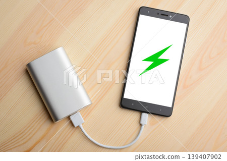 Modern smartphone lays on light wooden table while charging with power bank with quick charge. Illustration of green lightning is on the mobile phone screen 139407902