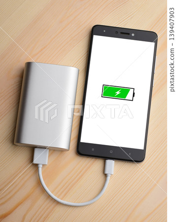 Modern smartphone lays on light wooden table while charging with power bank with quick charge. Illustration of green battery with lightning is on the mobile phone screen while charging Modern smartphone lays on light wooden table while charging with power bank with quick charge. Illustration of green battery with lightning is on the mobile phone screen while charging 139407903