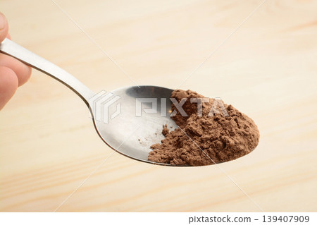 Hand holds spoon with cocoa powder above wooden table in kitchen Hand holds spoon with cocoa powder above wooden table in kitchen 139407909