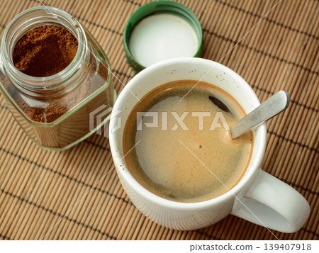 Photo of coffee in a white cup and spoon standing near the jar of coffee and green cover. Making aromatic coffee Photo of coffee in a white cup and spoon standing near the jar of coffee and green cover. Making aromatic coffee 139407918