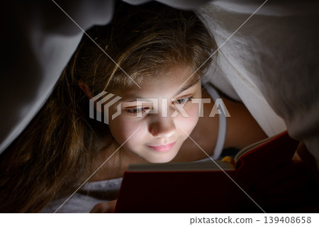 Little female child alone under the cover reading favourite fairy tales with a flashlight at night. Little female child alone under the cover reading favourite fairy tales with a flashlight at night. 139408658