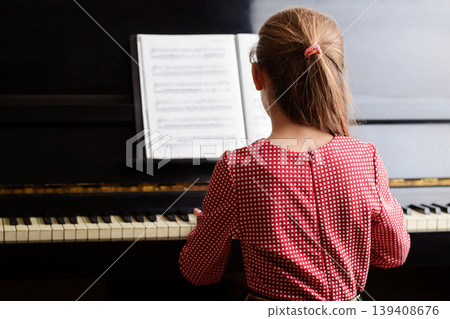 Little talented girl musician in a red dress playing the piano using folder with music sheet, performing beautiful classical music. Lifestyle concept. 139408676