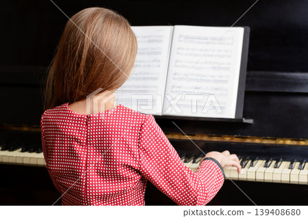 Small female child pianist in beautiful red dress sitting at piano with music sheet, playing classical music at the concert 139408680