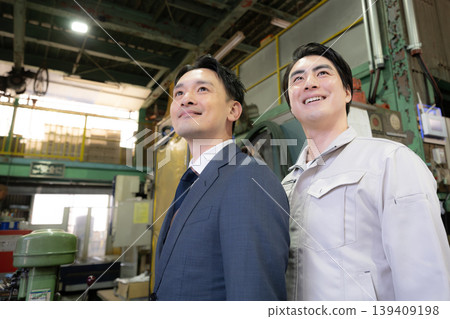 In a factory, a businessman and a worker, both smiling and looking up, gaze towards the future, demonstrating the trusting relationship between the two. In a factory, a businessman and a worker, both smiling and looking up, gaze towards the future, demonstrating the trusting relationship between the two. 139409198