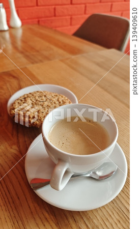 Coffee cup filled with espresso served on a white saucer alongside a round oatmeal cookie on a wooden table with a red brick wall in the background Coffee cup filled with espresso served on a white saucer alongside a round oatmeal cookie on a wooden table with a red brick wall in the background 139409610