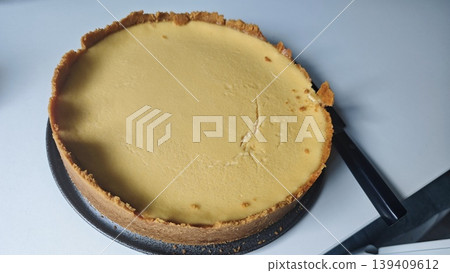 Round cheesecake with a golden crust placed on a white surface, a black knife positioned beside it ready for slicing, showcasing its smooth texture and creamy filling Round cheesecake with a golden crust placed on a white surface, a black knife positioned beside it ready for slicing, showcasing its smooth texture and creamy filling 139409612