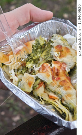Hand grips a foil tray containing a baked spinach and cheese pastry, revealing a golden crust and green filling, with an outdoor setting in the background 139409620