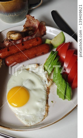 Breakfast plate featuring fried egg, sausages, bacon, mushrooms, cucumber, and tomato slices arranged on a white plate with a cup in the background Breakfast plate featuring fried egg, sausages, bacon, mushrooms, cucumber, and tomato slices arranged on a white plate with a cup in the background 139409621