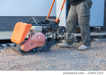 Worker uses a vibratory plate compactor for preparing a base layer. Essential step before paving, decking, or foundation installation. Worker uses a vibratory plate compactor for preparing a base layer. Essential step before paving, decking, or foundation installation. 139411263