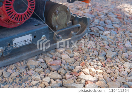 Close-up of a vibratory plate compactor working on a gravel base at a construction site. The machine is used for soil compaction and ground preparation before laying foundations or paving. Close-up of a vibratory plate compactor working on a gravel base at a construction site. The machine is used for soil compaction and ground preparation before laying foundations or paving. 139411264