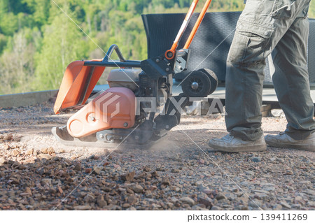 Plate compactor operating on dry gravel, creating a visible dust cloud. The image captures dynamic construction work and ground preparation. Plate compactor operating on dry gravel, creating a visible dust cloud. The image captures dynamic construction work and ground preparation. 139411269