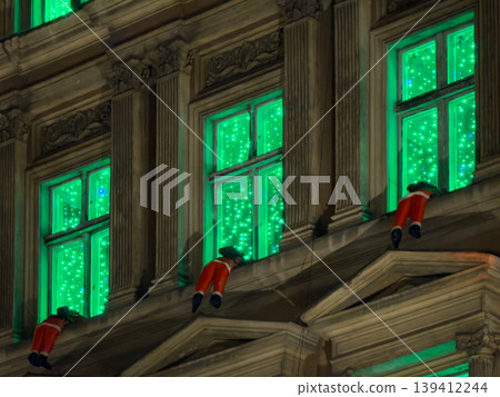 A historic building facade showcases vibrant green sparkling lights and whimsical red elf figures, creating a charming festive urban Christmas holiday decoration A historic building facade showcases vibrant green sparkling lights and whimsical red elf figures, creating a charming festive urban Christmas holiday decoration 139412244