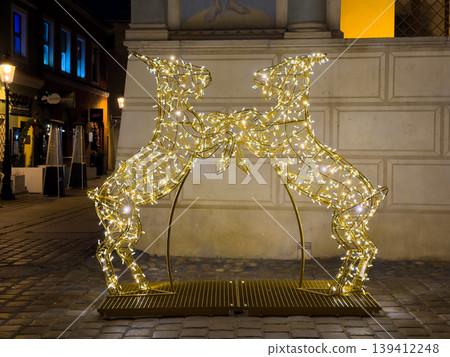 Two glowing golden deer light sculptures facing each other, creating a captivating festive display for holiday season celebration on a cobblestone city street at night Two glowing golden deer light sculptures facing each other, creating a captivating festive display for holiday season celebration on a cobblestone city street at night 139412248