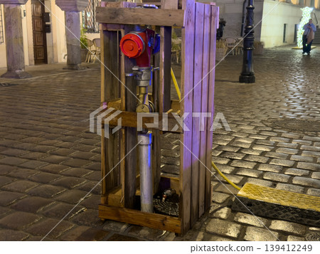 Red fire hydrant securely protected inside a wooden crate on a wet cobblestone street at night, symbolizing urban infrastructure, temporary work, and emergency safety 139412249