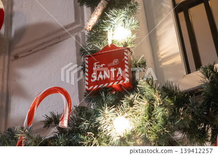 A festive red 'For Santa' mailbox ornament hangs from a sparkling Christmas garland with glowing lights, a candy cane, embodying holiday spirit and childhood wonder A festive red 'For Santa' mailbox ornament hangs from a sparkling Christmas garland with glowing lights, a candy cane, embodying holiday spirit and childhood wonder 139412257