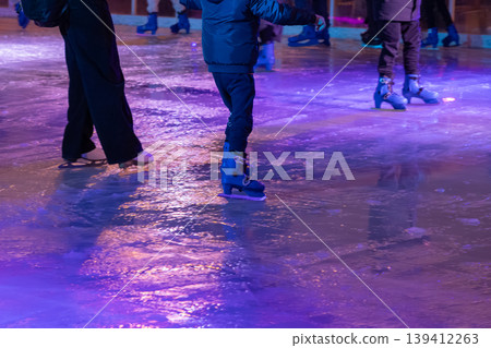Ice skaters enjoying an evening session on a vibrant rink illuminated with glowing purple and blue light, reflecting festive winter recreation and holiday joy Ice skaters enjoying an evening session on a vibrant rink illuminated with glowing purple and blue light, reflecting festive winter recreation and holiday joy 139412263