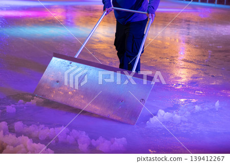 Person cleaning a brightly lit ice rink with a large metal scraper, preparing the pristine surface for exciting winter sports and festive community events 139412267