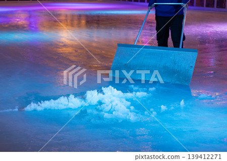 Worker pushing an ice scraper, removing slush from a vibrantly lit ice rink, ensuring a perfectly smooth surface for public winter skating Worker pushing an ice scraper, removing slush from a vibrantly lit ice rink, ensuring a perfectly smooth surface for public winter skating 139412271