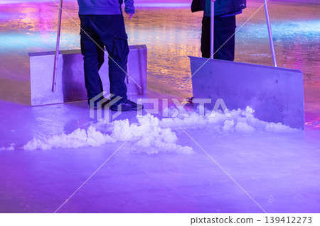 Maintenance workers scrape an ice rink, pushing snow piles under vibrant purple and blue lights, reflecting a lively winter sports and leisure event Maintenance workers scrape an ice rink, pushing snow piles under vibrant purple and blue lights, reflecting a lively winter sports and leisure event 139412273