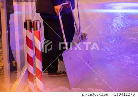 Worker clearing ice and snow from a public rink with a large shovel under vibrant purple lighting, maintaining the surface for winter sports and activities 139412279