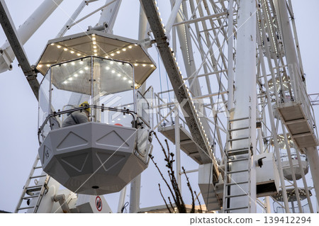 Illuminated white Ferris wheel with a passenger enjoying a ride in a numbered cabin, symbolizing amusement park fun and exciting outdoor leisure activity Illuminated white Ferris wheel with a passenger enjoying a ride in a numbered cabin, symbolizing amusement park fun and exciting outdoor leisure activity 139412294