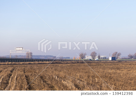 Harvested field next to highway with overhead sign and distant industrial buildings, illustrating rural transition and transportation infrastructure 139412398