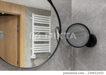 Modern bathroom featuring a large round mirror reflecting a towel warmer and a magnifying mirror, providing comfort and functionality for personal hygiene Modern bathroom featuring a large round mirror reflecting a towel warmer and a magnifying mirror, providing comfort and functionality for personal hygiene 139412433