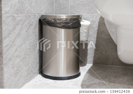 Sleek stainless steel waste bin with a black liner, positioned in a modern tiled public restroom to maintain excellent hygiene and cleanliness standards 139412439