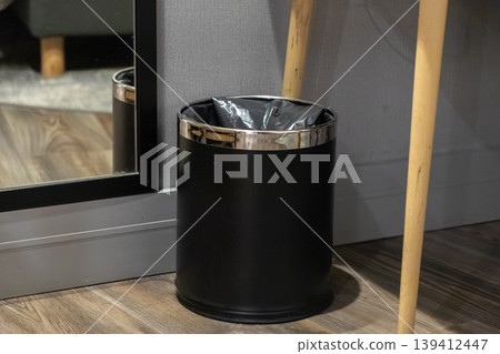 Modern black waste bin with metallic rim and a trash bag on a wooden floor, symbolizing cleanliness and efficient waste management in an interior 139412447