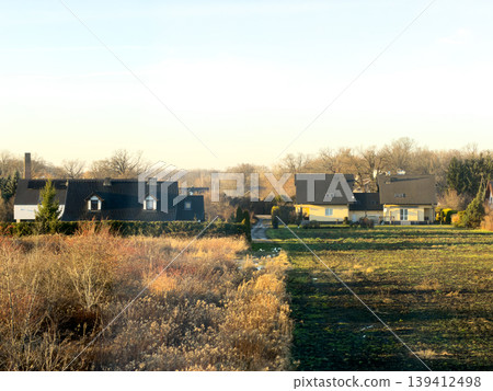 Suburban houses overlooking contrasting fields of dry autumn vegetation and vibrant green land under a bright clear sky, representing peaceful coexistence and seasonal landscape Suburban houses overlooking contrasting fields of dry autumn vegetation and vibrant green land under a bright clear sky, representing peaceful coexistence and seasonal landscape 139412498