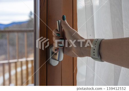 Female hand with elegant green nail polish opening a modern balcony door handle, revealing a scenic mountain view, suggesting fresh air and peaceful domestic comfort Female hand with elegant green nail polish opening a modern balcony door handle, revealing a scenic mountain view, suggesting fresh air and peaceful domestic comfort 139412552