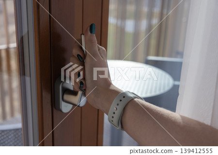Close-up of a woman's hand with a smartwatch opening a modern wooden balcony door, symbolizing security, access, and the comfort of home or hotel accommodation Close-up of a woman's hand with a smartwatch opening a modern wooden balcony door, symbolizing security, access, and the comfort of home or hotel accommodation 139412555