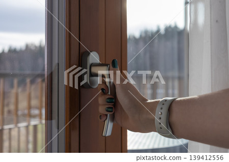 Woman's hand opens a modern window handle, welcoming fresh air into the home and connecting with the serene outdoor natural landscape 139412556
