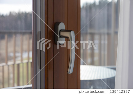 Modern window handle on a brown wooden frame with a blurred balcony view, representing home comfort, security, and a connection to nature Modern window handle on a brown wooden frame with a blurred balcony view, representing home comfort, security, and a connection to nature 139412557
