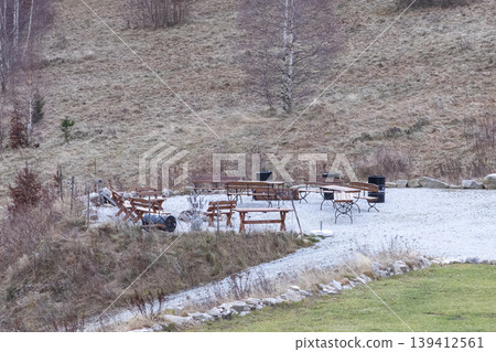Rustic outdoor dining area with wooden tables, benches, and barbecue pits on a gravel terrace, nestled in a dry, natural hillside Rustic outdoor dining area with wooden tables, benches, and barbecue pits on a gravel terrace, nestled in a dry, natural hillside 139412561