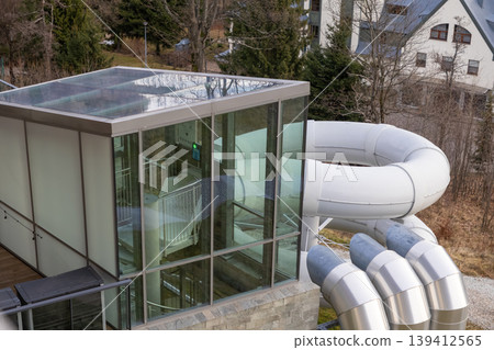 Modern water slide structure with white curved tubes and a glass entrance, offering fun recreational activity at an outdoor resort destination Modern water slide structure with white curved tubes and a glass entrance, offering fun recreational activity at an outdoor resort destination 139412565